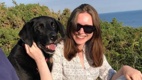 Gareth Williams Rhiannon has long dark hair and is wearing a white top and sunglasses. She sits next to the family's black labrador, Jet, near Langland in the summer of 2022.