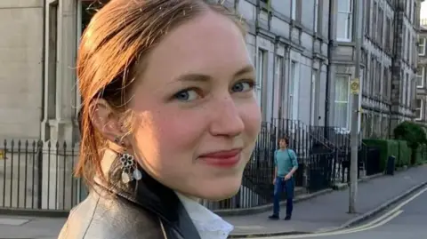 Cordelia Ostler Rhiannon has brown hair, tied back and wears a black leather jacket and white blouse. He turns to the camera and smiles. Behind him is a row of houses in Edinburgh where he studied at university.